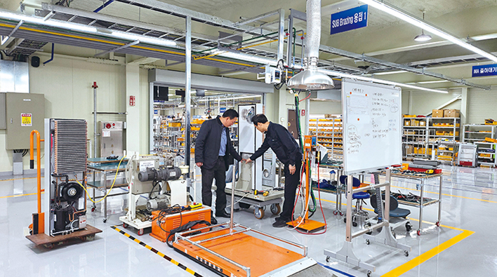 Two workers inspecting automated equipment on the manufacturing floor of a smart factory. Various machines, a whiteboard, and piping systems are arranged together.