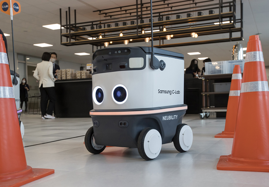 삼성 C-Lab과 NEUBILITY 로고가 표시된 자율주행 로봇이 실내에서 주황색 안전콘 사이를 주행 중인 모습. 주변에는 스타트업 전시 또는 행사장 분위기의 사람들이 있음